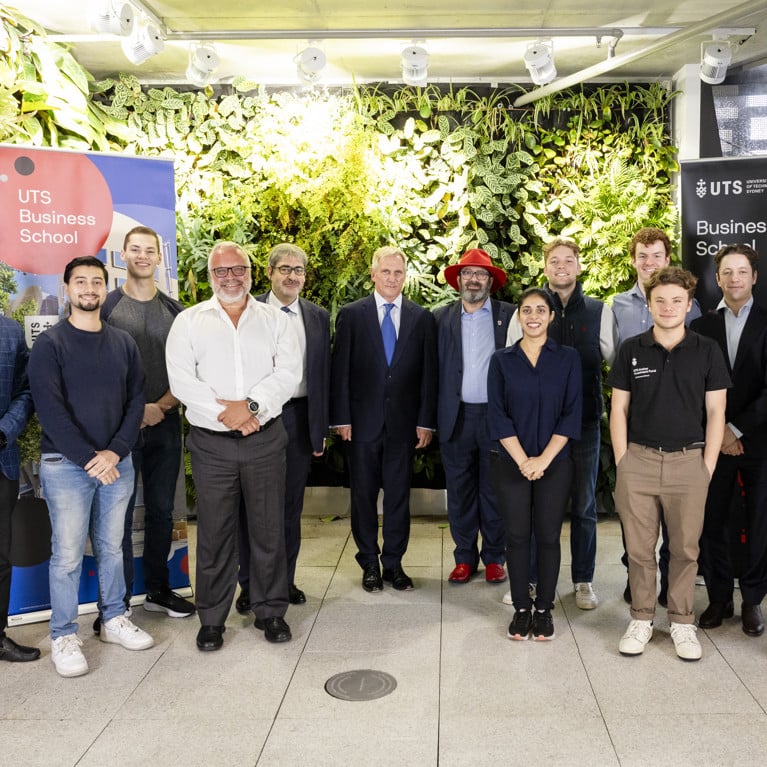 Academics, students and industry guest lecturers are standing together at the UTS Business School event, photographed in front of a lush green living wall with UTS Business School banners on display on either side of the group. They are facing the camera and smiling shoulder to shoulder in a line.