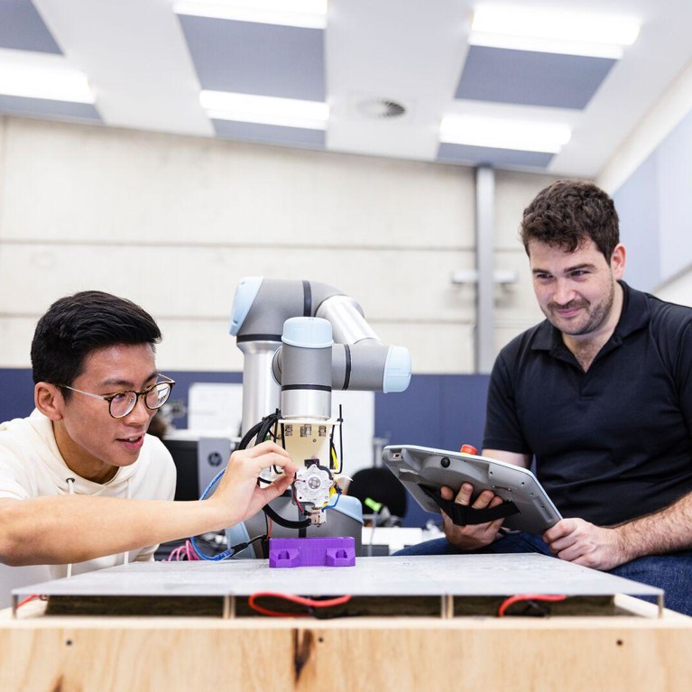 Man wearing glasses adjusts a robotic arm and man holding tablet watches.