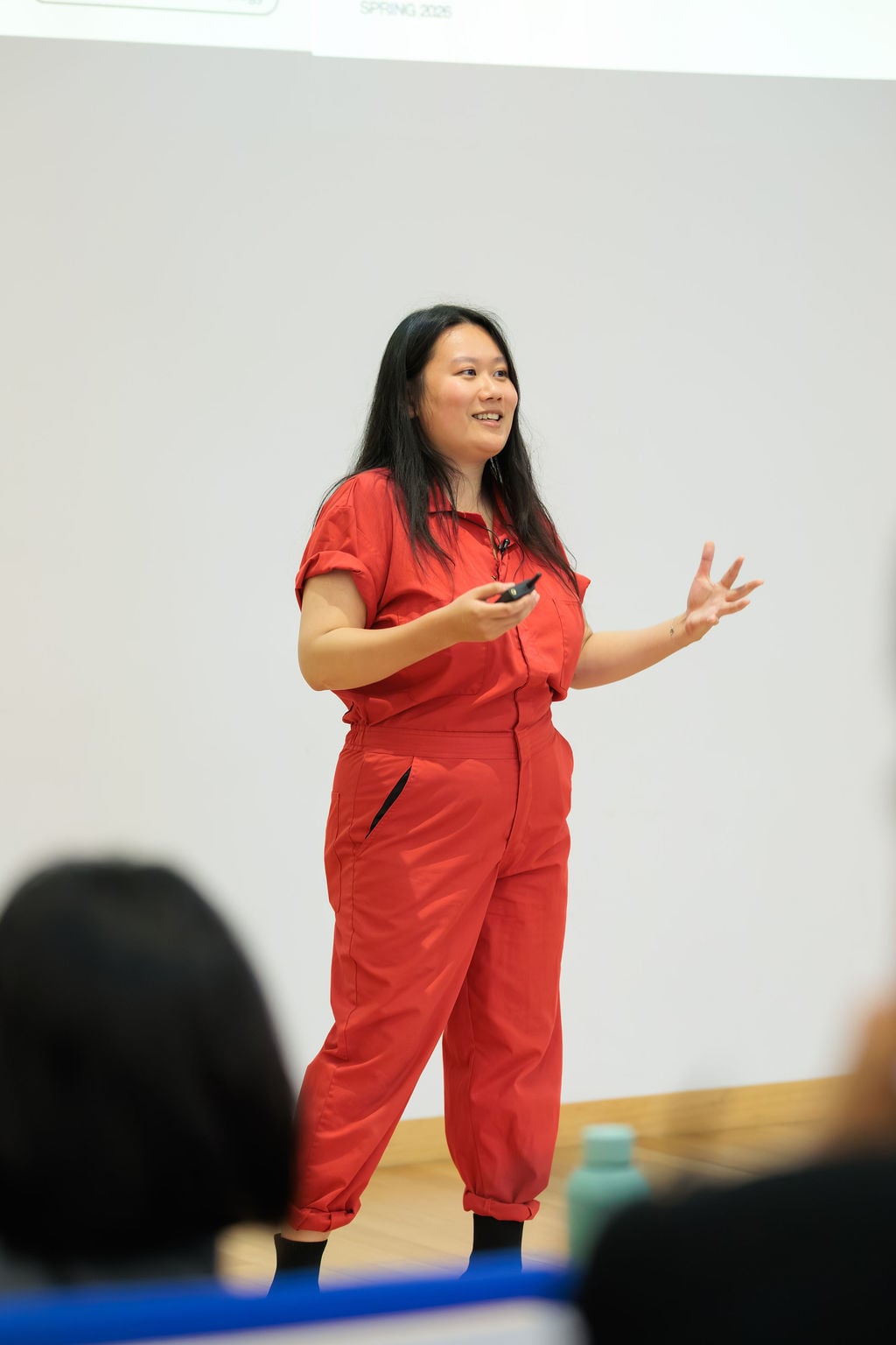 Shuya Gong speaking at the front of the auditorium wearing a red jumpsuit
