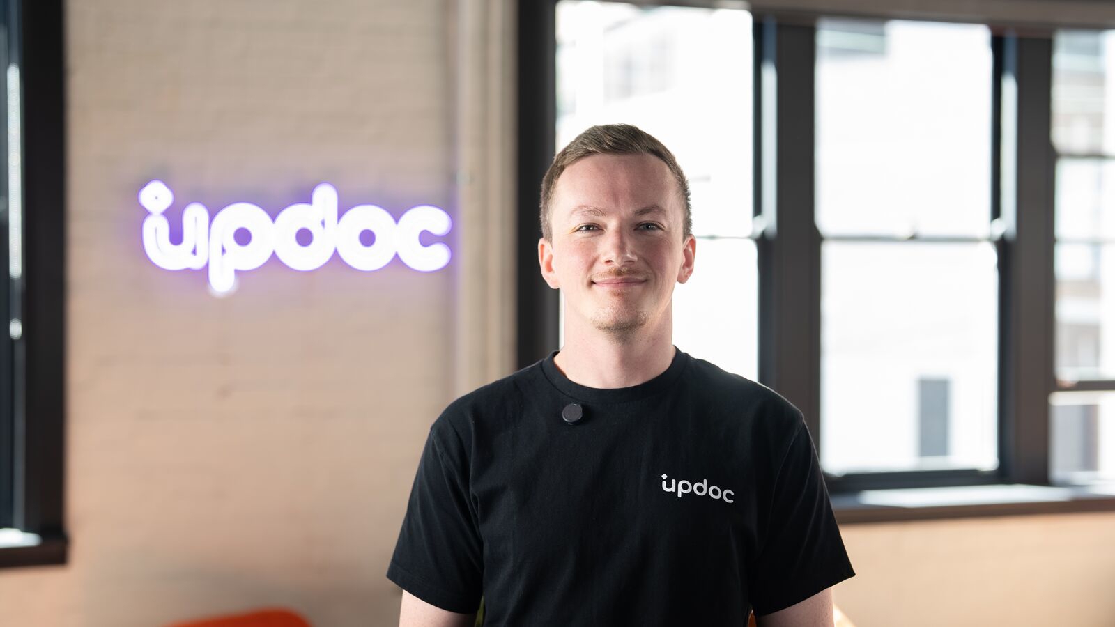 Updoc co-founder and BIT Alumni Dylan Coyne in a black t-shirt standing in the Updoc office. There is a neon purple 'Updoc' sign behind him.