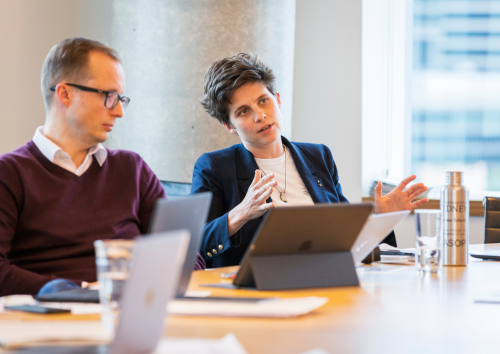 Two individuals engaged in discussion at a conference table, laptops open. One gestures expressively, conveying an analytical tone in a modern office setting.