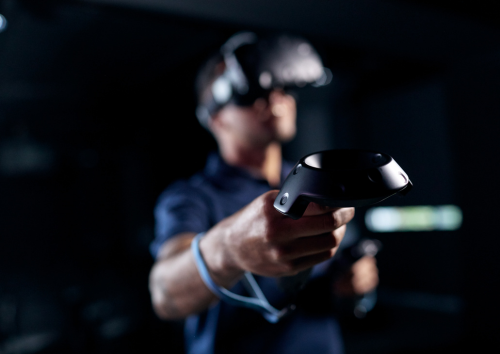 A person wearing a virtual reality headset holds and points a VR controller forward, with a dark, blurred background.