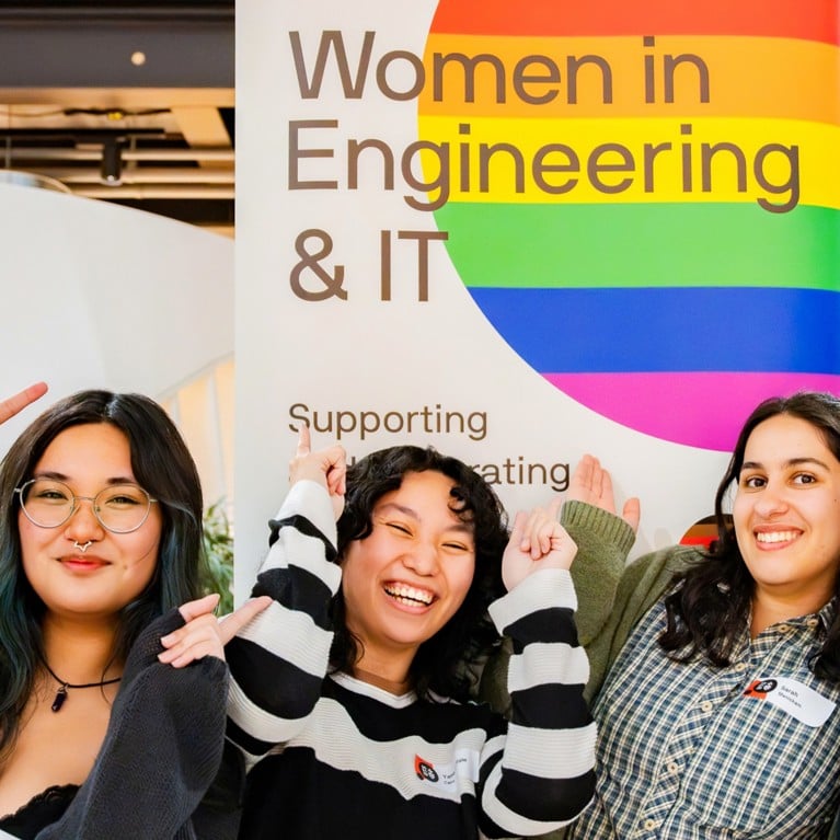 People standing in front of a pride rainbow banner for Women in Engineering and IT