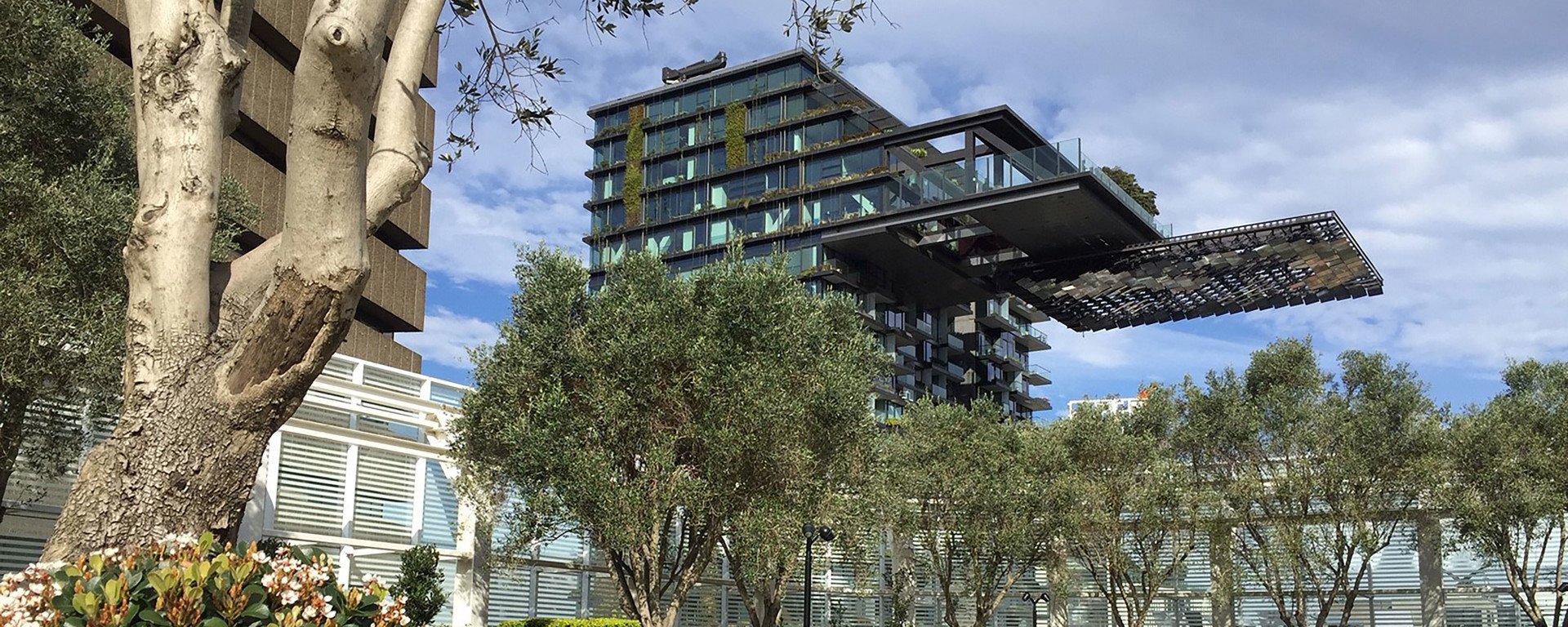 A modern glass building with greenery on balconies, set against a cloudy sky. Foreground features trees and flowers, creating a natural contrast.