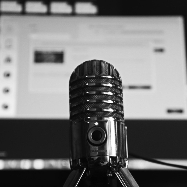 Black and white image with a microphone in the foreground and a desktop computer screen in the background.