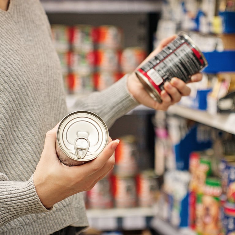 A woman choosing canned food at a store, reading the label. By Pavel Adobe Stock