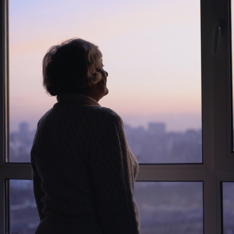 A senior woman looking out a high-rise building window silhouetted against a sunrise or sunset. Picture by Synthex/Adobe Stock