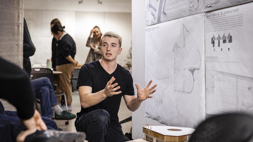 A young man gestures animatedly while presenting architectural plans on a wall. Several people in the background listen and take notes, creating a collaborative and focused atmosphere.