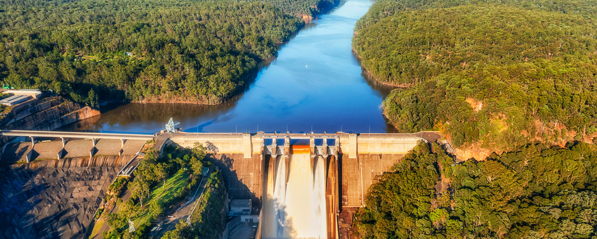 Aerial view of a dam releasing water into a river surrounded by dense, green forests. The clear blue sky and calm atmosphere convey tranquility and majesty.