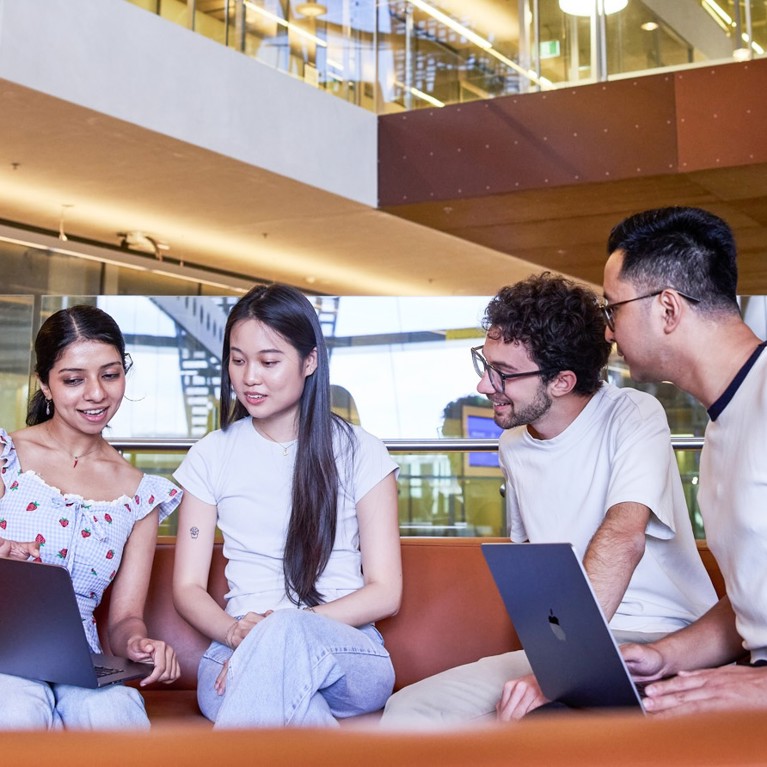 Four students sit on a curved orange couch with laptops, engaged in discussion. Modern, well-lit library setting, exuding collaboration and focus.