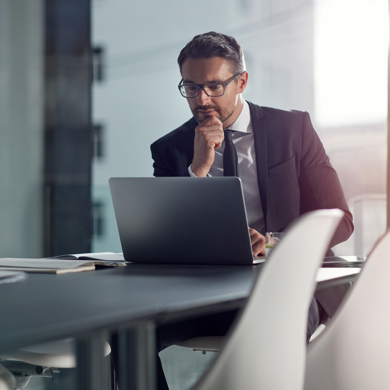 A businessman in a white shirt and a black suit and tie works at a laptop in an office environment