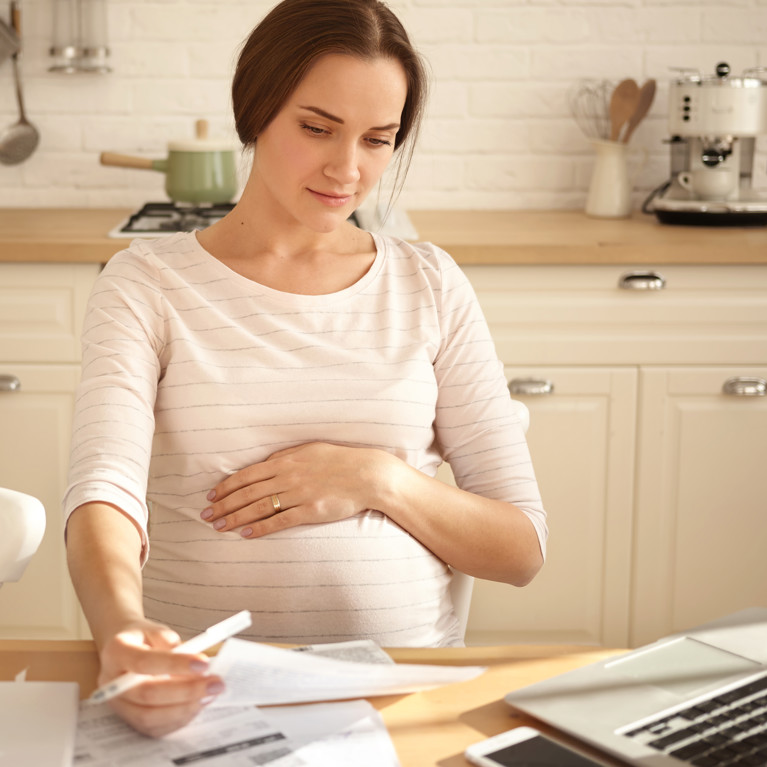 Indoor shot of young pregnant woman keeping one hand on belly, holding sheet of paper, paying bills online using laptop computer. Picture by shurkin_son/Adobe Stock