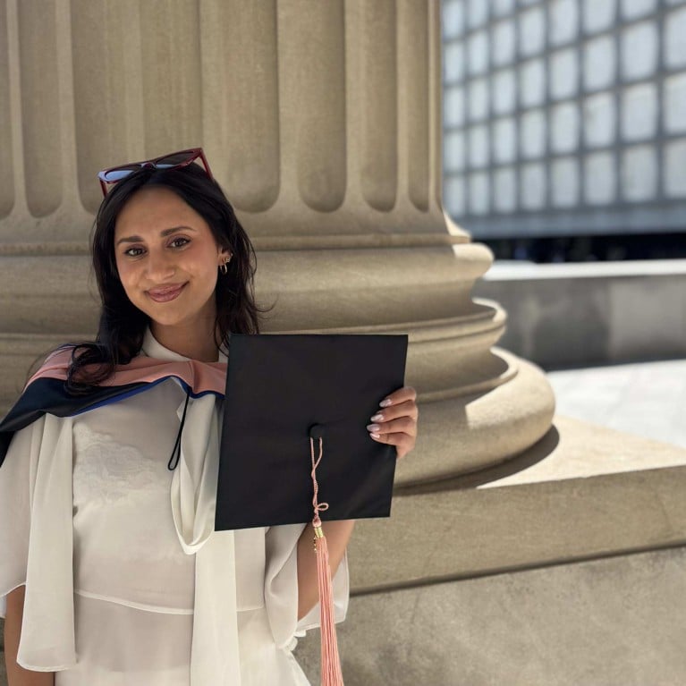Jaya at her graduation. She wears a white dress and her graduation outfit and stands in front of a stone column