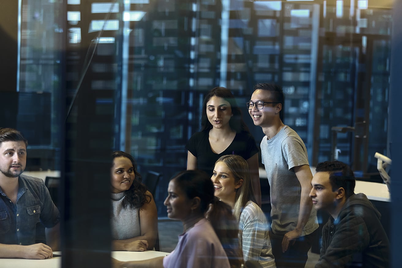 A group of people collaborating around a table in a modern office, viewed through a glass wall.