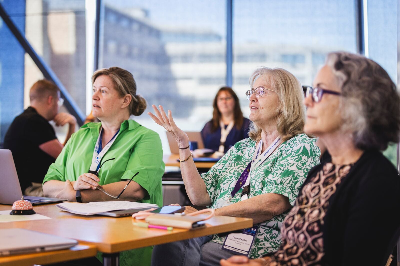 Participants seated around a table during a workshop, listening to a speaker and contributing to discussion, with laptops, notebooks and conference lanyards visible.