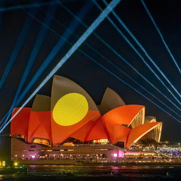 The Sydney Opera House is illuminated at night with vibrant red and yellow Indigenous flag art projections. Blue laser beams radiate into the night sky.