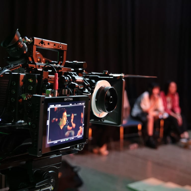 Close-up of a professional video camera on a film set, focused on two people in a dimly lit room. The atmosphere is creative and focused.