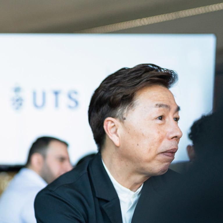 Three men engaged in discussion in a modern setting, with one looking attentive. A UTS logo is visible on a screen in the background.