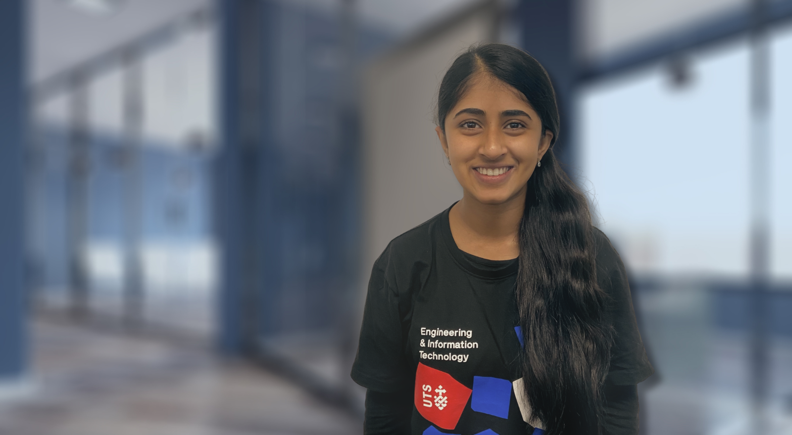 Aalia in an Engineering and IT t-shirt standing in front of a blurred classroom background