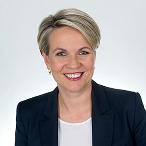 Tanya Plibersek appears in the foreground wearing a navy blazer over a white top, posed against a clean studio-style background. She has short, light‑coloured hair styled neatly, with a professional, front‑facing posture.