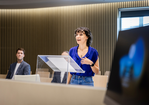 A woman stands at a podium, presenting to an audience seated in rows, engaged and attentive.