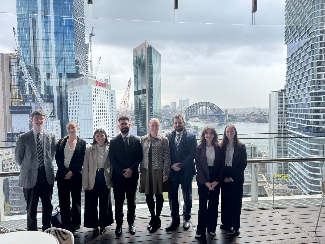 Jordan Chambers, Annabelle Macclelland, Ahmad Hamad, Chiara D'Ambrosio, Challita Tahhan, Charlotte Antoinette, Sophie Hambling, Thomas Fleming stand on a balcony with Sydney harbour in the background.