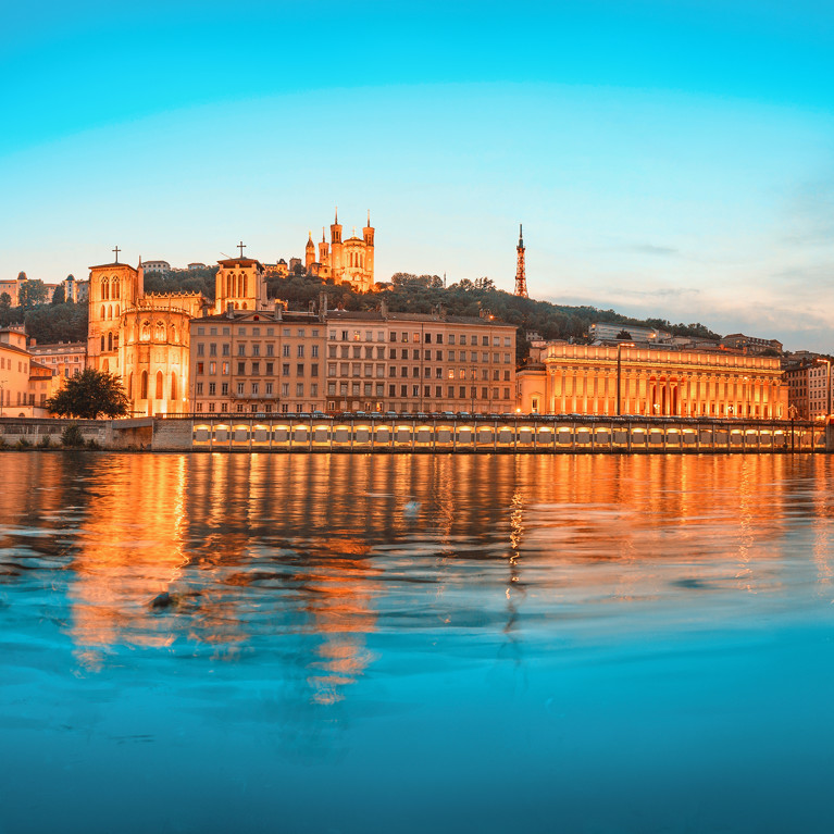 Illuminated cityscape of Lyon at dusk, featuring historic buildings on the waterfront and a modern suspension bridge, reflected in the calm river.