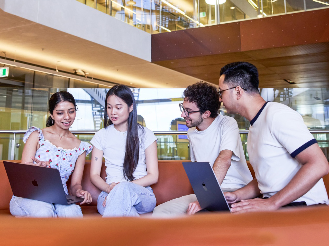 Four students sit on a curved orange couch with laptops, engaged in discussion. Modern, well-lit library setting, exuding collaboration and focus.