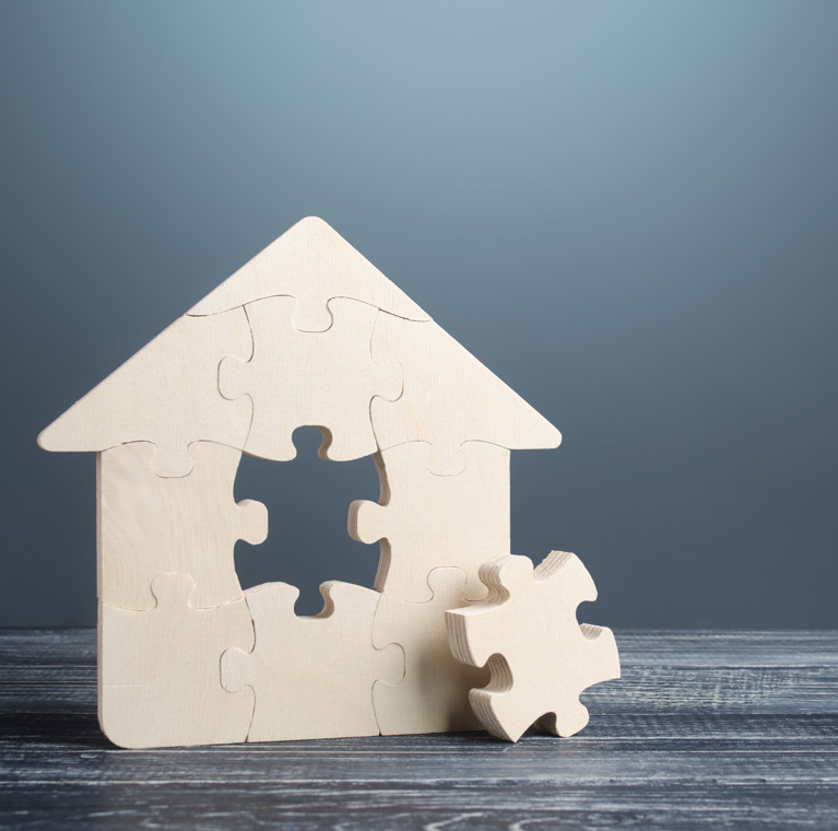 A wooden house sits on a table made out of jigsaw peices, one of the pieces at the centre has been plucked out and is laying beside the house on the table.