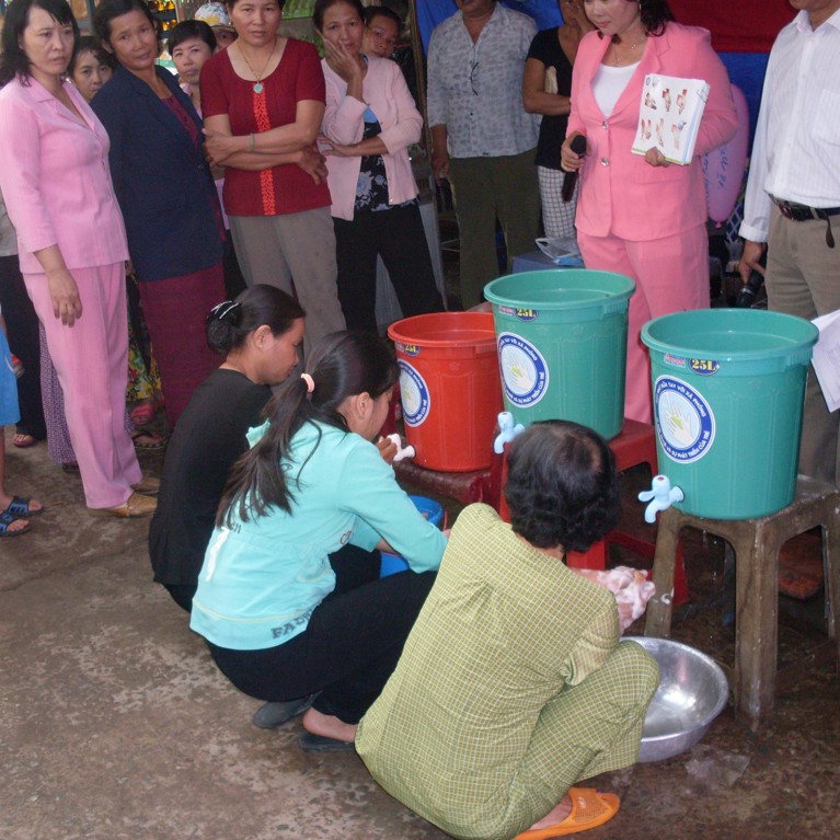 People crouching washing hands from buckets with taps and onlookers surrounding