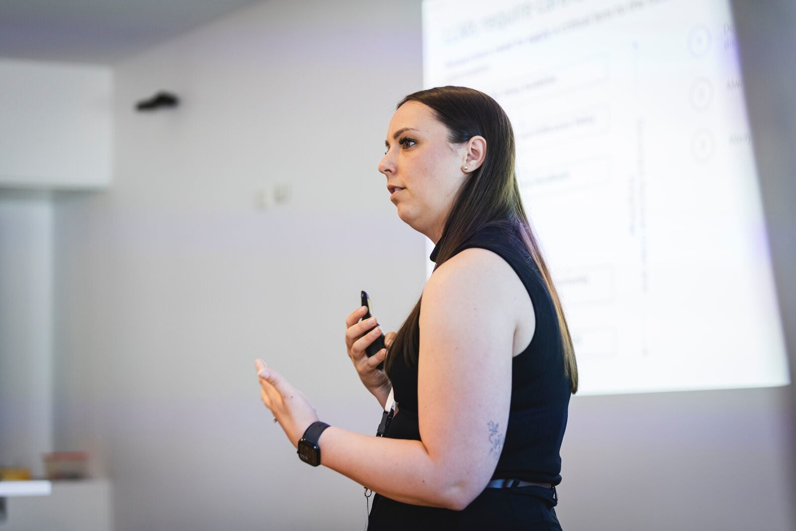 In the foreground, a female speaker in a black dress is gesticulating in one hand and holding a buzzer in the other as she addresses an audience we cannot see. Behind her in the background is a PowerPoint presentation whose slides projected onto the wall.