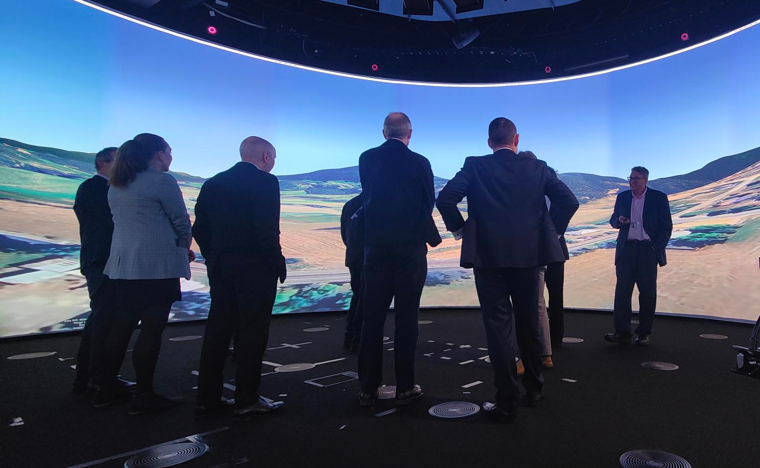 A group of people stand in a dark room with a large, curved digital screen displaying a vast landscape. The mood is focused and collaborative.