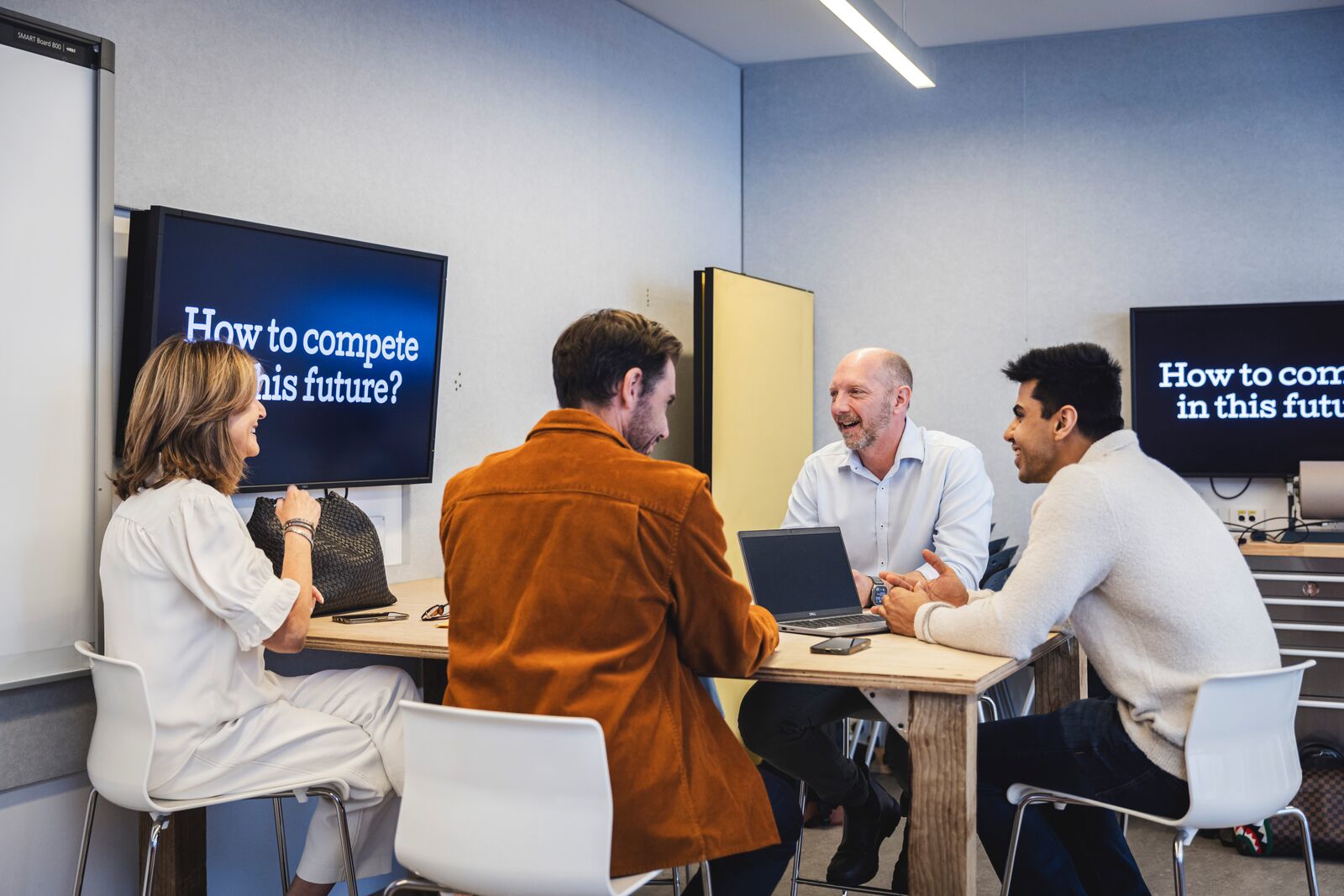 A group of co-workers sit around a table with their laptops, they are in discussion. Behind them on screens is a prompt that reads "How do we compete in this future?".