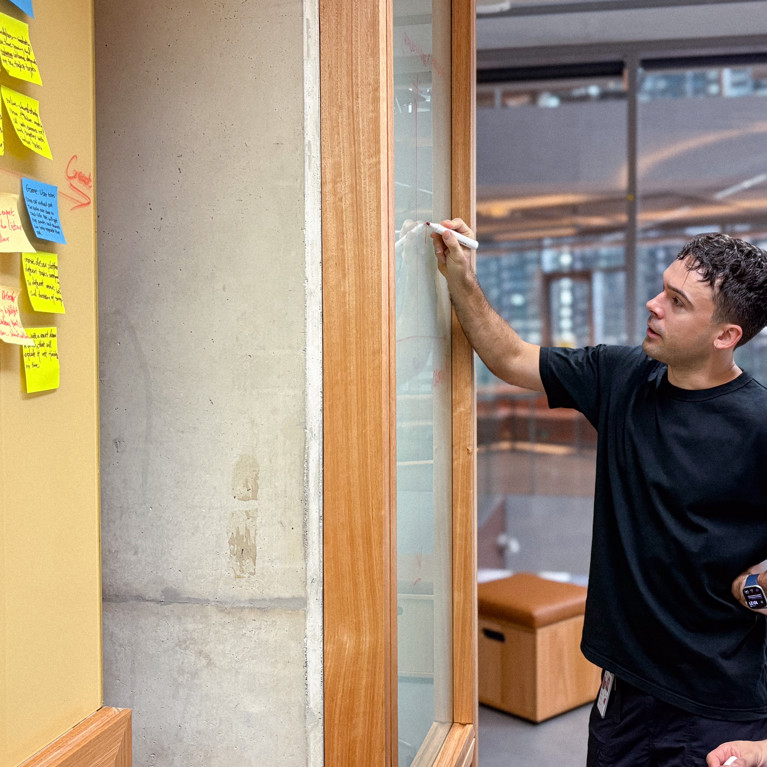 Two men in black shirts collaborate in an office, writing on a glass board. A wall with colourful sticky notes is on the left. The scene is focused and innovative.
