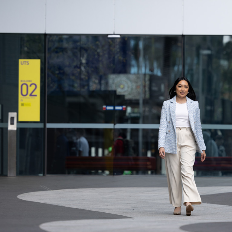 Arhita, wearing a light blue blazer and beige trousers, strides forward outside UTS building 2