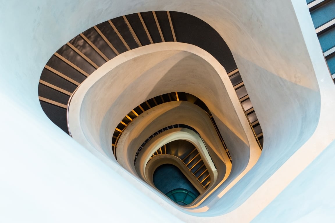 View from above of a spiraling staircase with smooth, white walls and dark steps, creating an abstract, modern design with a sense of depth and flow.