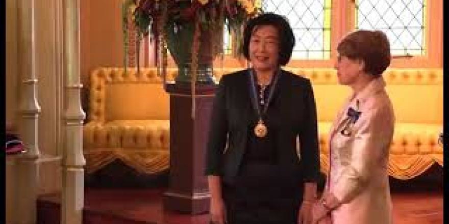 Medium range shot of NSW Governor Margaret Beazley placing Jie Lu's investiture insignia medal around her neck. The Governor is wearing a light pink satin dress suit, her shoes are black pumps with a sparkly silver block heel. Jie Lu is wearing a knee length dress with thick block stripes in the colours black, dark green and navy blue. She has a matching green tailored jacket. She is wearing black suede heels with a pointed toe. Jie Lu's face is pictured smiling at the Governor.