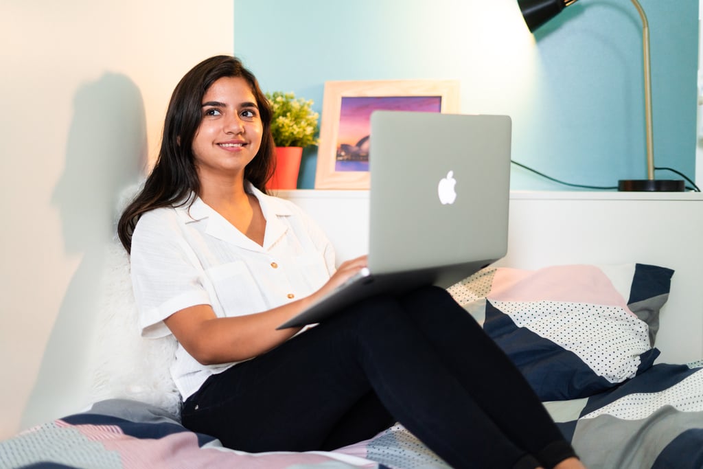 Young woman on laptop in bed, smiling while looking out the window.