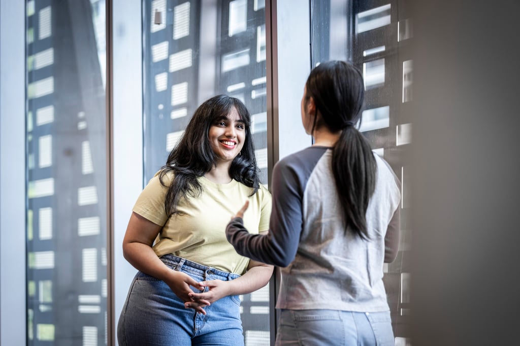 Two women converse by large windows. One woman, smiling, leans against a wall; the other faces her, gesturing.