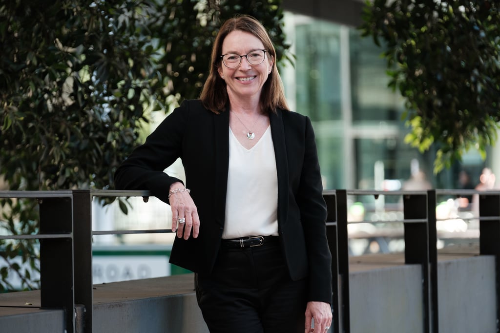 In the foreground Rowena stands to face the viewer in a black blazer, white shirt and black pants. She's wearing glasses and smiling at the viewer and she leans on a railing at UTS. In the background is Building 8 and trees.