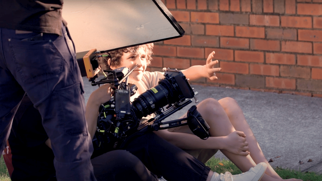A child being photographed while sitting on grass beside a concrete path and a brick wall.