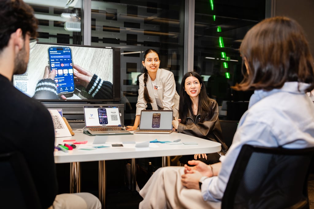 2 Tech Fest participants presenting their project to 2 attendees. Attendees are seated facing the presenters who are standing next to a digital screen