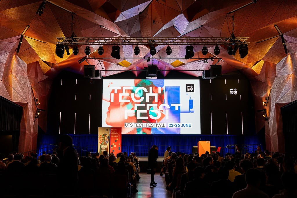 Image taken from back of crowd  towards stage in UTS Great Hall. On stage is a speaker at a lectern with a Tech Fest digital banner behind