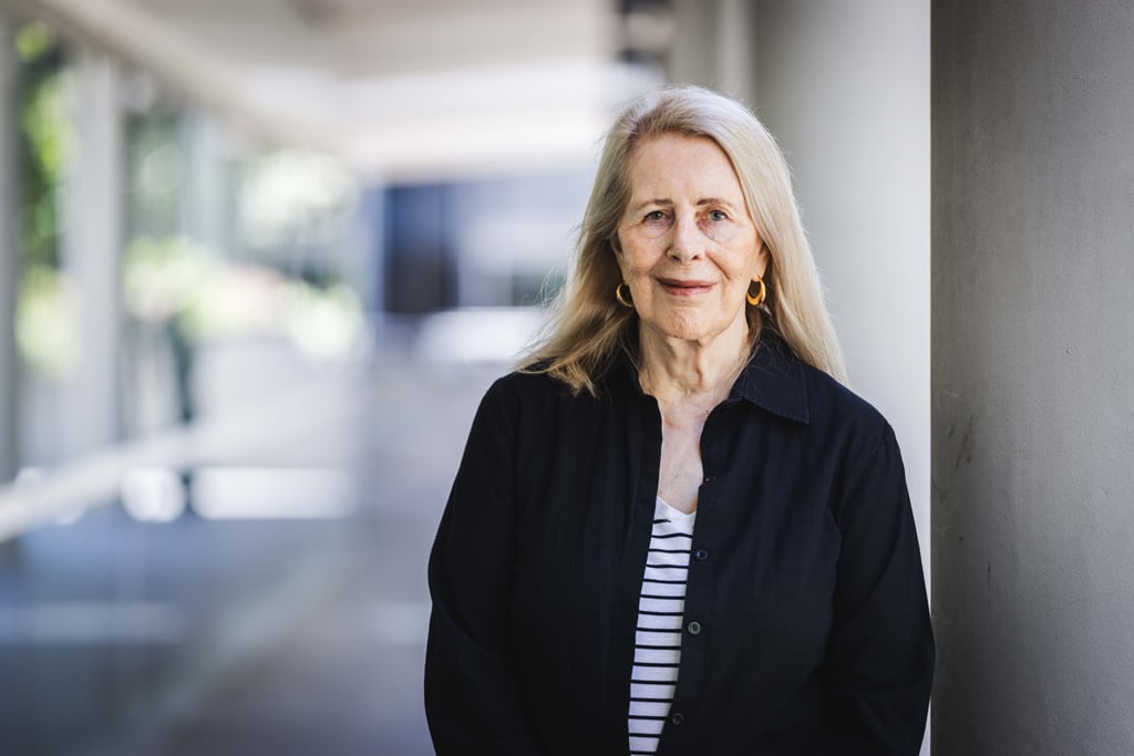 Anne Summers stands in the foreground next to a concrete pillar. She's wearing a black jacket and white top with blue stripes and gold earnings. The background of the UTS campus walkway is blurred with pillars and foliage.