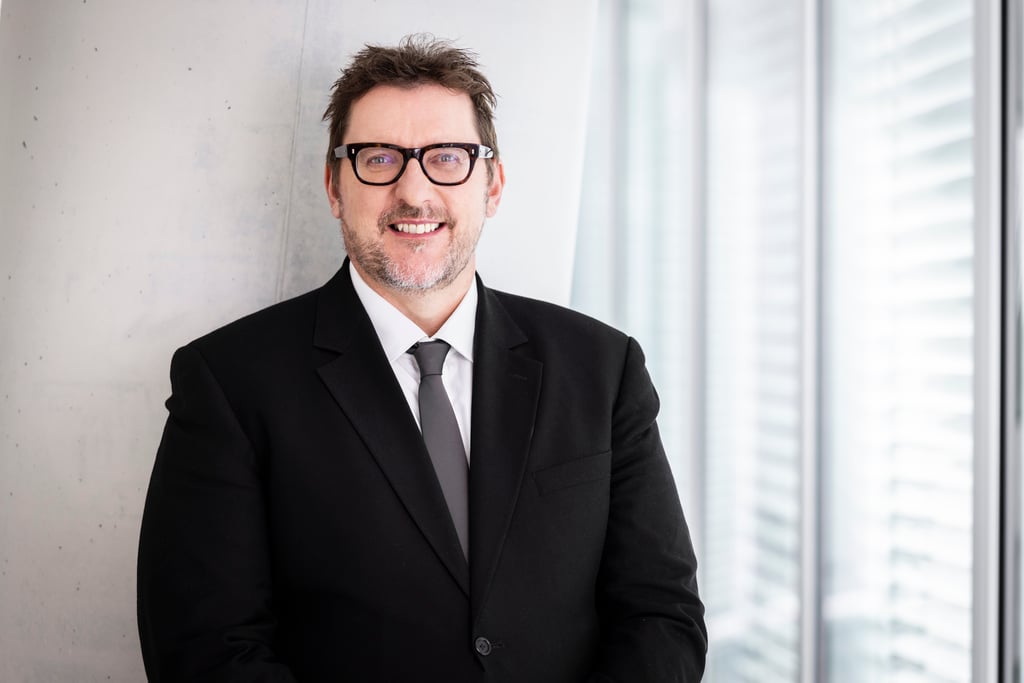 Carl stands smiling at the viewer in a black suit with black rimmed glasses, standing in front of a concrete pillar in Building 8.