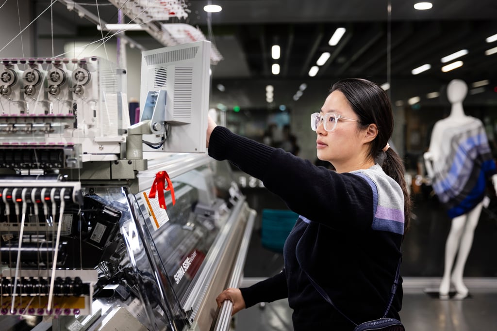 A woman in glasses operates a knitting machine in a UTS studio. She looks focused, with a mannequin draped in fabric in the background.