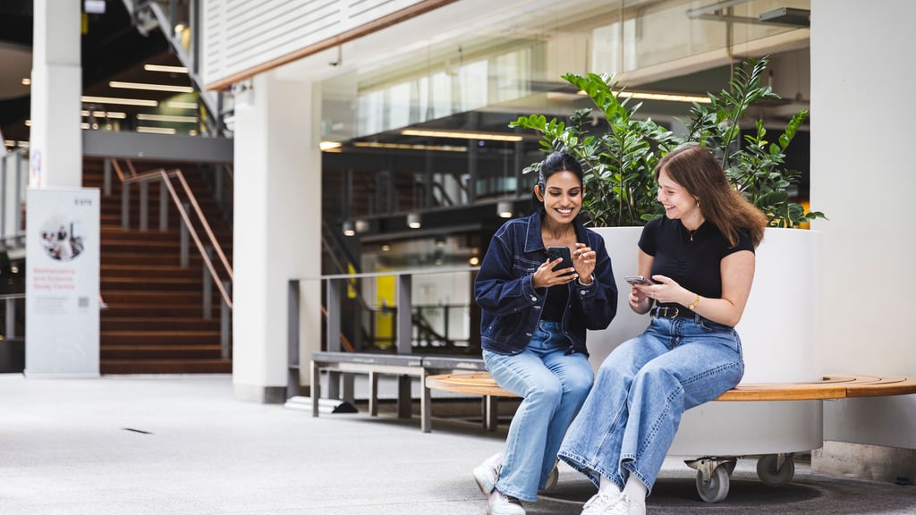 Two female students sitting on a bench looking at phones.