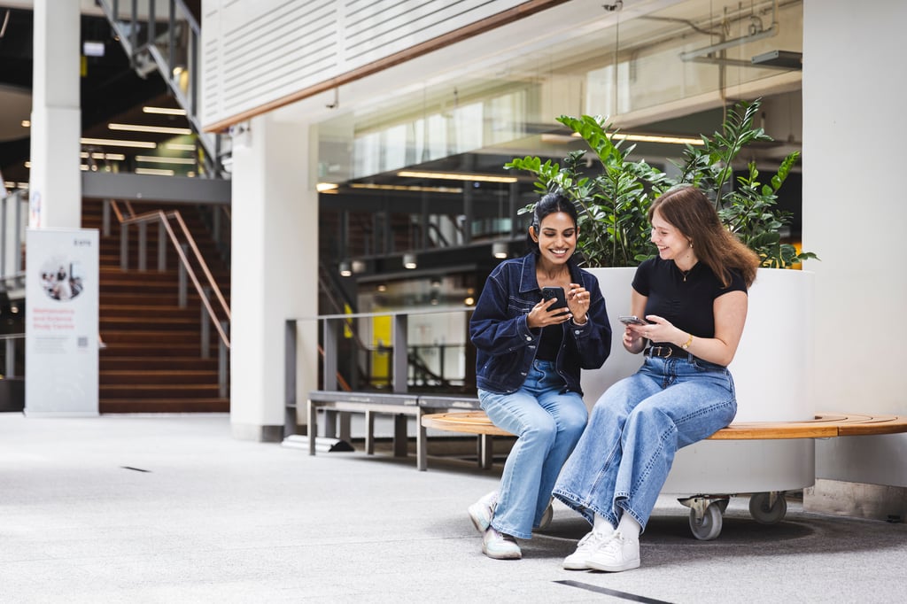 Two female students sitting on a bench looking at phones and smiling.