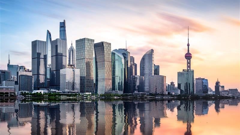 A skyline of modern skyscrapers reflects on a calm river during sunset.