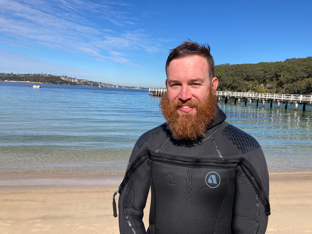 Portrait of Brennan wearing a wetsuit, standing on the beach with the shoreline behind him.